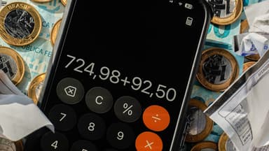 Calculadora de celular somando valores sobre cédulas e moedas de real, ao lado de comprovantes amassados