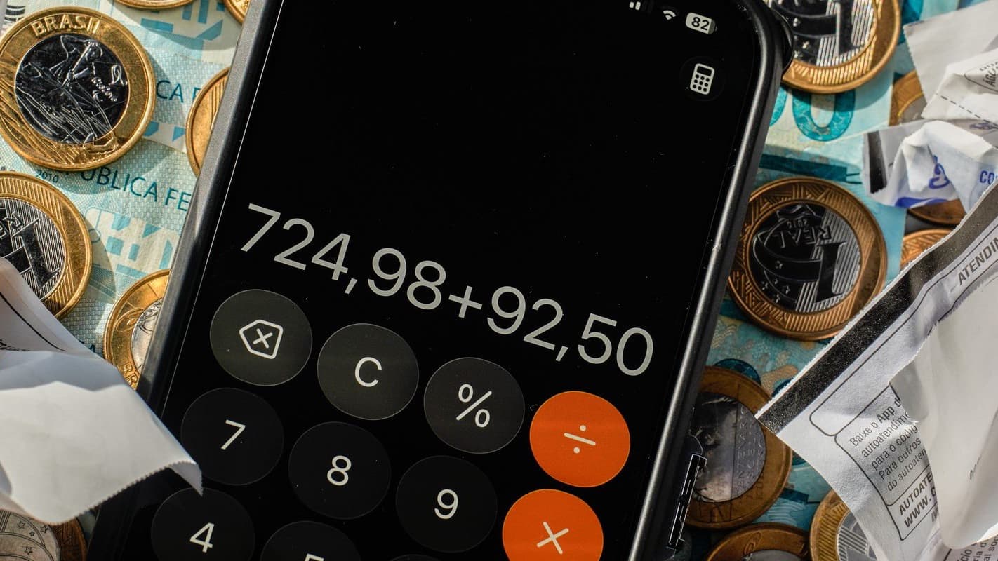 Calculadora de celular somando valores sobre cédulas e moedas de real, ao lado de comprovantes amassados