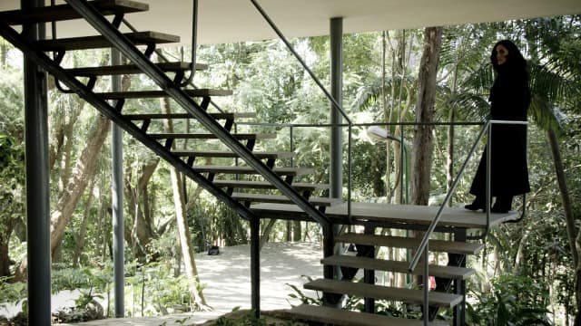 Escadaria externa de metal e madeira da Casa de Vidro de Lina Bo Bardi, cercada por vegetação, com pessoa de preto no topo