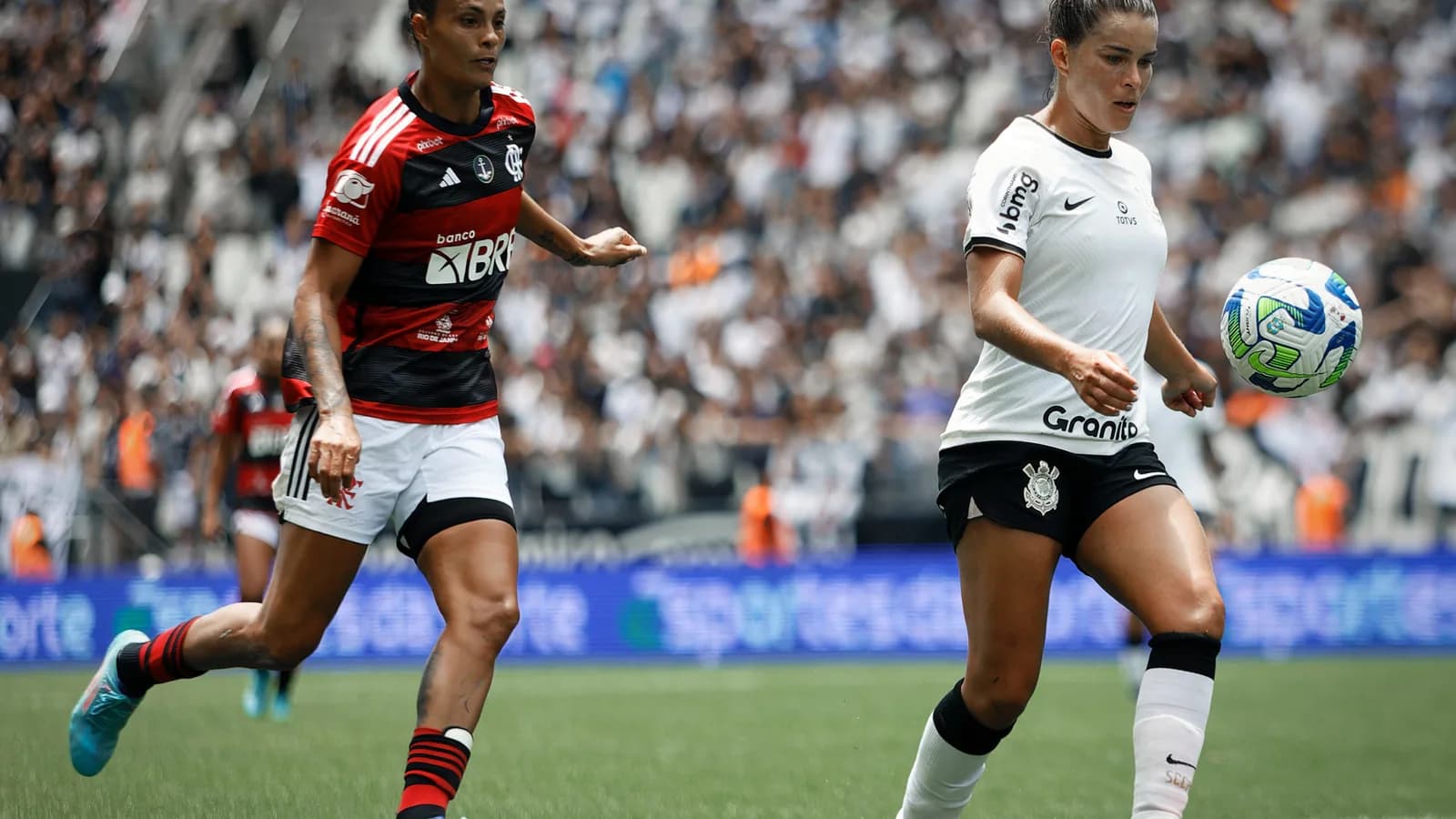 Jogadoras do Corinthians e do Flamengo disputam a bola em partida de futebol feminino, com a torcida ao fundo