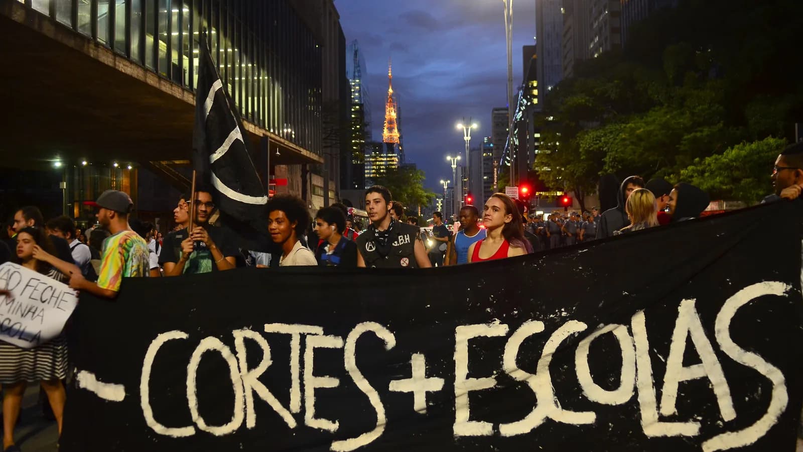 Estudantes em manifestação à noite, segurando faixa com os dizeres 'menos cortes, mais escolas' (foto de arquivo)
