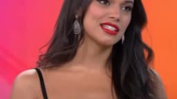 Alane Dias sorri em entrevista de televisão, com fundo colorido em tons de rosa e laranja