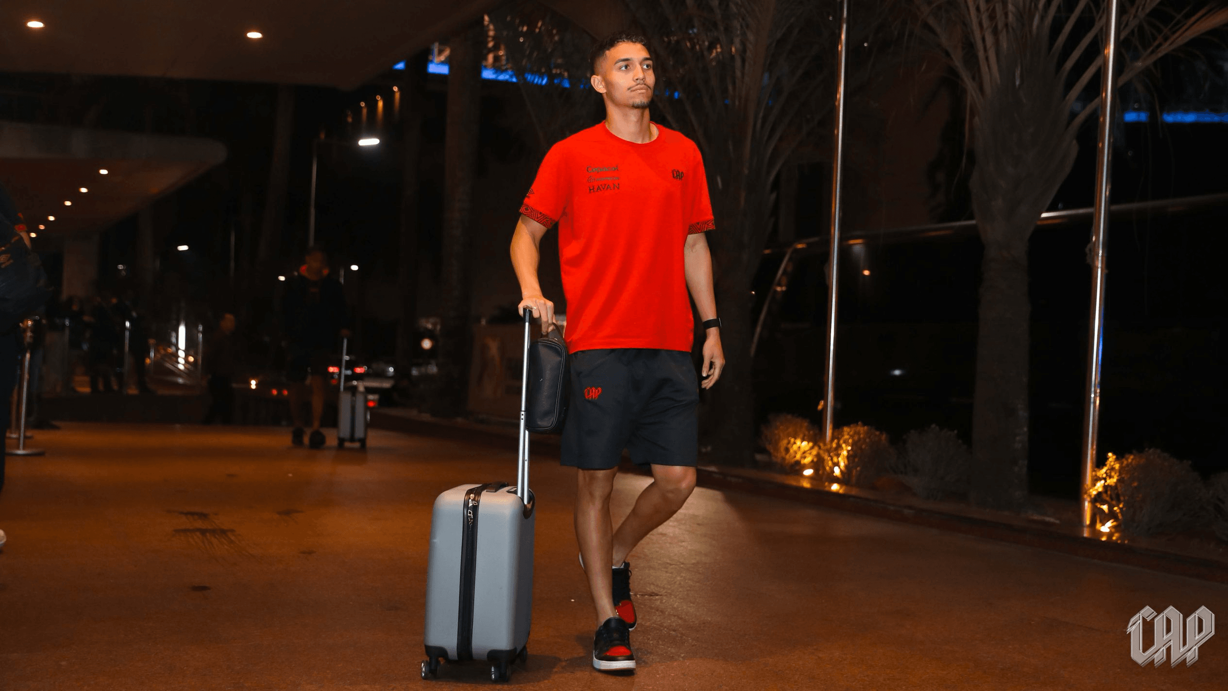 Jogador do Athletico Paranaense com uniforme de viagem do clube caminha com mala em hotel, na véspera do jogo contra o Santos