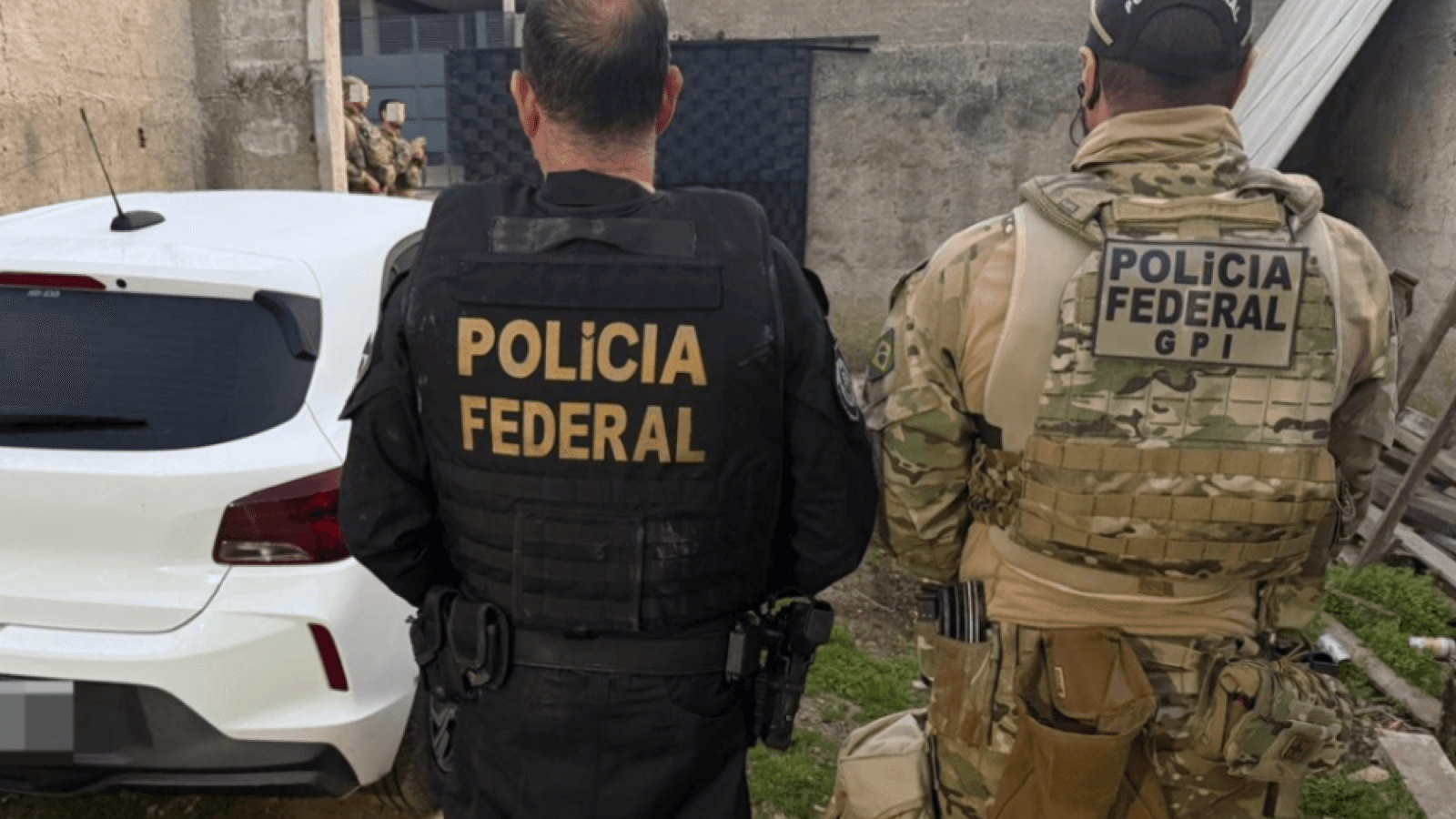 Dois agentes da Polícia Federal, de costas, ao lado de viatura durante operação contra tráfico de armas