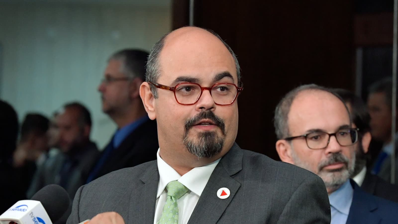 Mateus Simões, de terno cinza e gravata verde, fala a jornalistas em entrevista coletiva; ao fundo, homens de terno acompanham