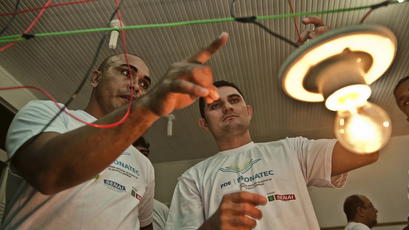 Dois trabalhadores em treinamento de qualificação profissional mexem em instalação elétrica