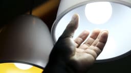 Mão de uma pessoa diante de duas luminárias acesas, uma com luz amarela e outra com luz branca; imagem de arquivo sobre consumo de energia elétrica