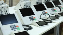 Fileira de urnas eletrônicas da Justiça Eleitoral sobre mesas, em preparação para eleição