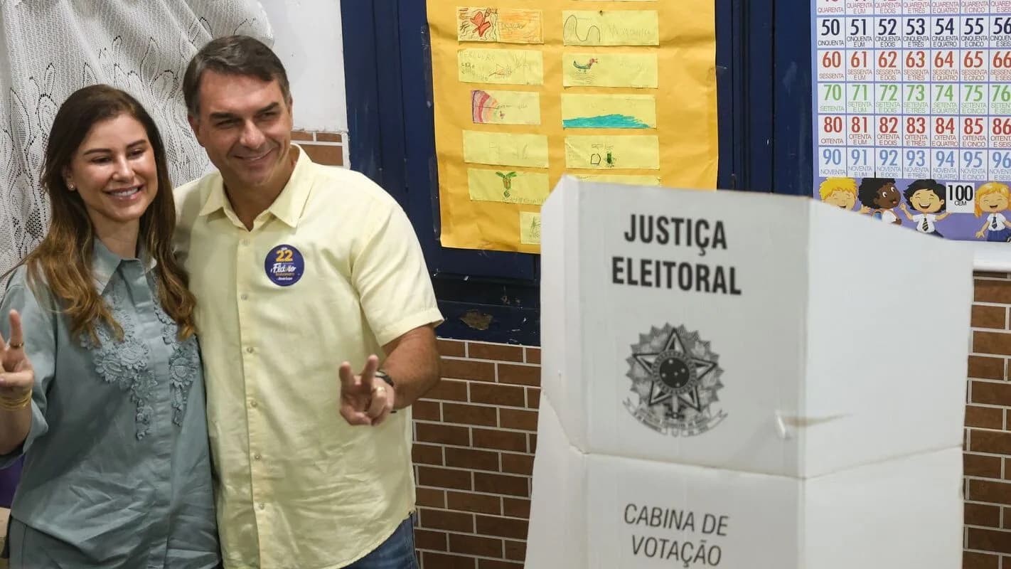 Flávio Bolsonaro e Fernanda Bolsonaro fazem sinal de positivo ao lado de urna eletrônica em seção eleitoral