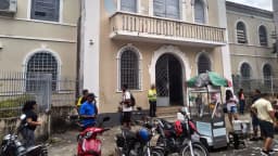 Eleitores em fila do lado de fora de um local de votação, com motos estacionadas e mesários organizando o acesso