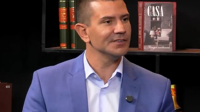 Douglas Ruas, de terno azul, fala em entrevista com estante de livros ao fundo