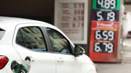Carro branco é abastecido em posto de combustível com placas de preço da gasolina ao fundo