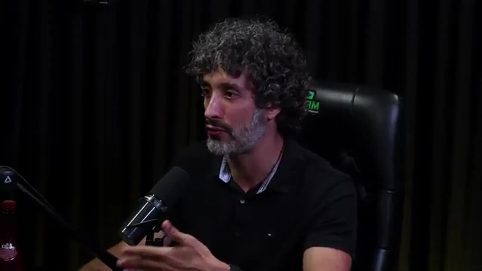 Carlos Eduardo Grimaldi, de cabelo cacheado grisalho e barba, gesticula com a mão enquanto fala ao microfone em estúdio de podcast com cortina escura ao fundo