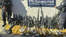 Dois policiais militares do Rio de Janeiro perfilados junto a fuzis e tabletes de drogas apreendidos, em frente a faixa da Polícia Militar do Estado do Rio de Janeiro