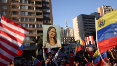 Manifestante segura retrato pintado de María Corina Machado em meio a bandeiras dos Estados Unidos e da Venezuela em manifestação de apoio à opositora