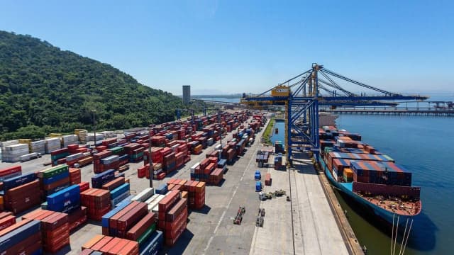 Contêineres empilhados em porto, com navio cargueiro atracado ao fundo
