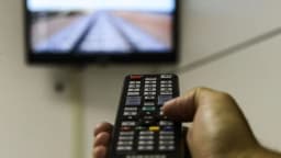 Mão segura controle remoto em frente a uma TV