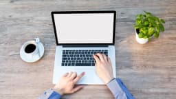 Xícara de café ao lado de um notebook em uma mesa de madeira, com mãos digitando no teclado