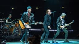 Banda U2 se apresenta ao vivo no palco durante turnê, com Bono no vocal e os demais integrantes tocando