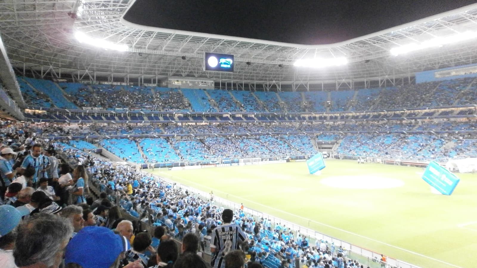 Torcida do Grêmio lota a Arena em jogo anterior à noite, com o gramado ao fundo