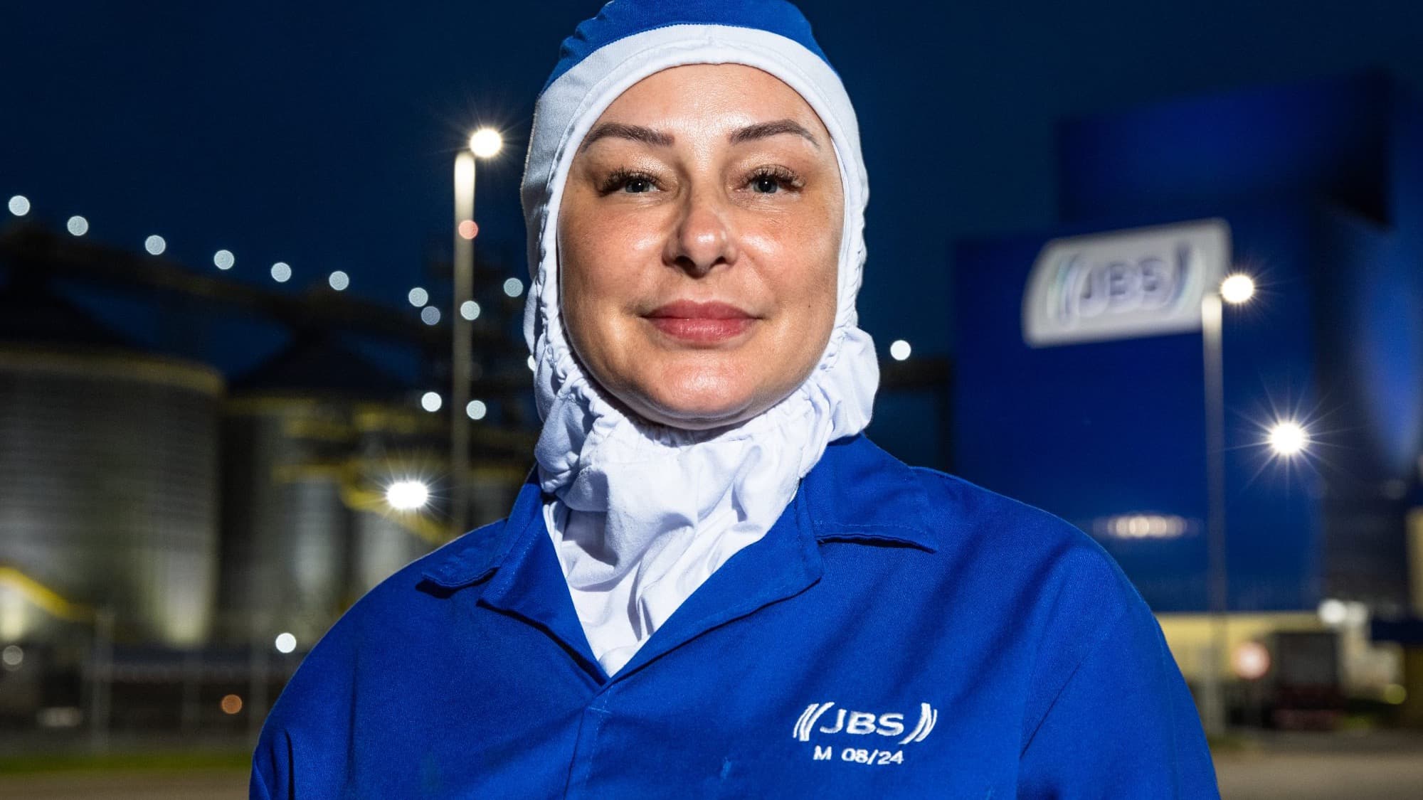 Colaboradora da JBS em frente a unidade da empresa durante a noite, com logo da marca ao fundo