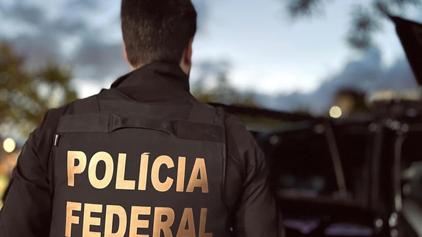 Agente da Polícia Federal visto de costas, em operação ao lado de um veículo, com o colete escrito Polícia Federal