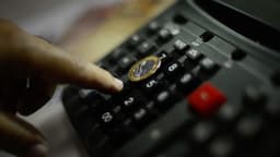 Mão apertando o teclado de uma calculadora com uma moeda de um real apoiada sobre as teclas