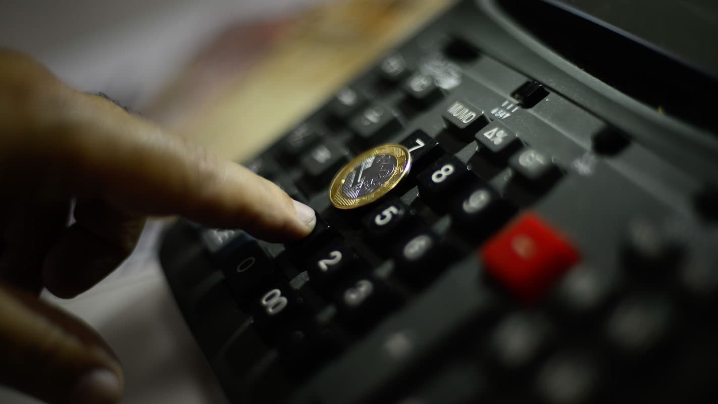 Mão apertando o teclado de uma calculadora com uma moeda de um real apoiada sobre as teclas