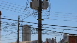 Poste de rede elétrica com transformadores e fiação em área urbana, prédios ao fundo (foto de arquivo, contexto)