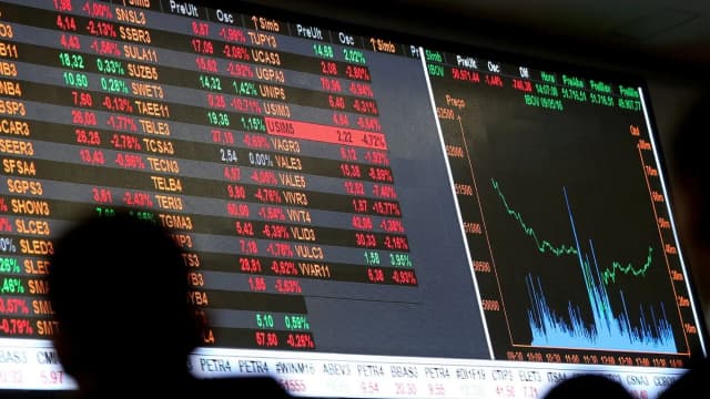 Painel eletrônico exibe cotações de ações e o índice Ibovespa, com pessoa observando os dados