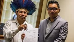 Integrante da Apib com cocar entrega documento de representação a autoridade pública, ao lado de outro representante da entidade, em imagem de arquivo