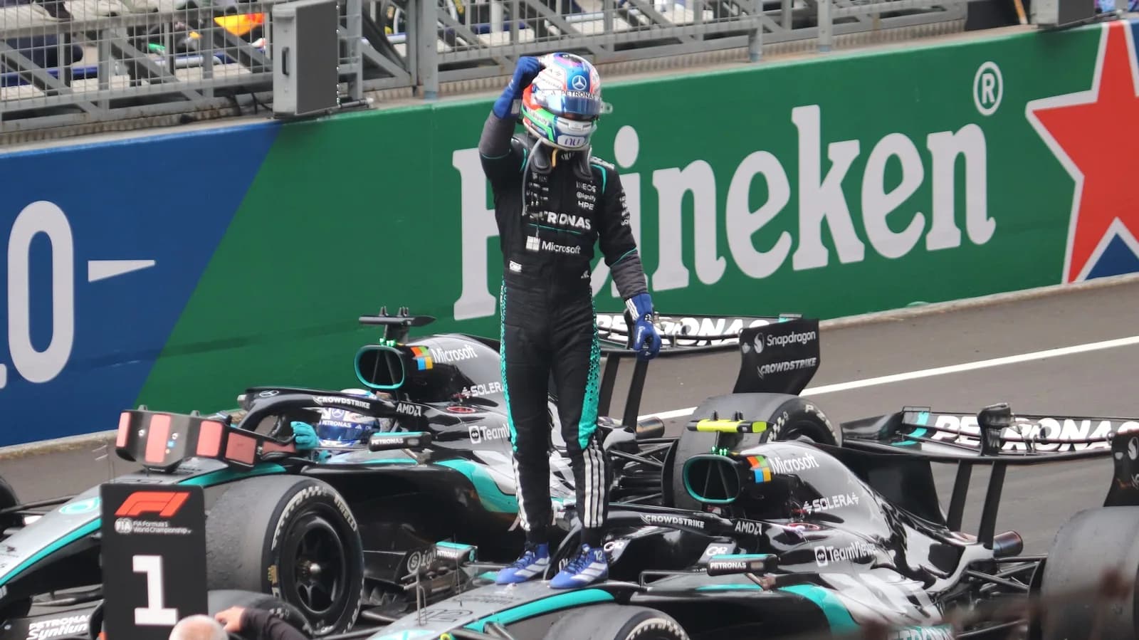 Piloto de macacão preto da Mercedes em pé sobre o carro de Fórmula 1, comemorando vitória, com bandeira do Heineken ao fundo