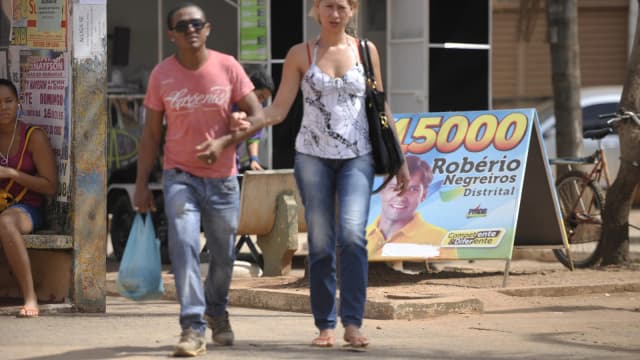 Pedestres caminham por calçada com placa de propaganda eleitoral ao fundo