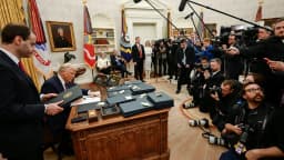 Donald Trump assina documentos em sua mesa no Salão Oval, cercado por fotógrafos e assessores