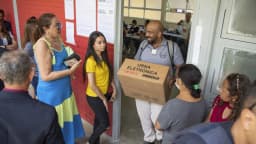 Eleitores em fila e mesário carregando caixa de urna eletrônica em local de votação em Brasília, eleições de 2022