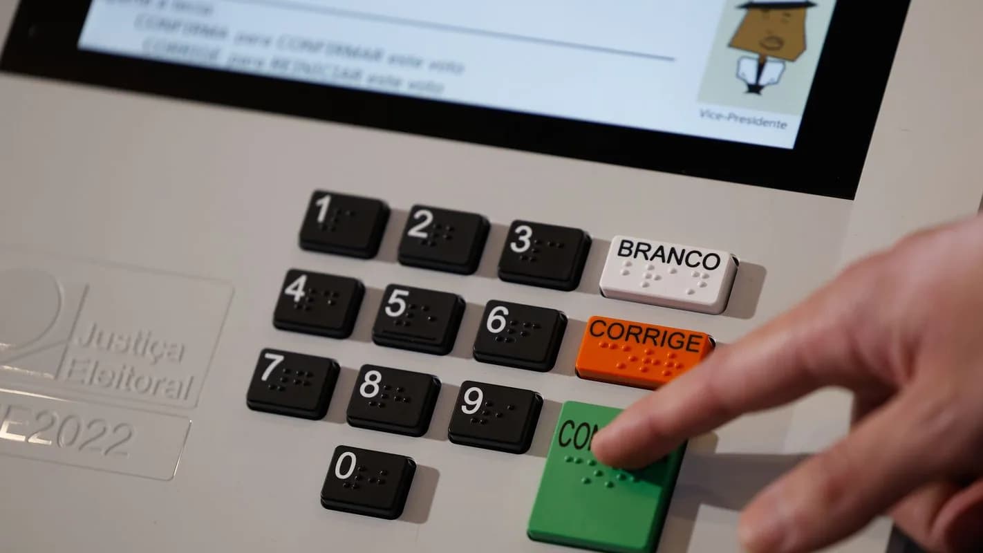 Dedo pressiona tecla verde em braile de urna eletrônica, ao lado de teclas numéricas também em braile