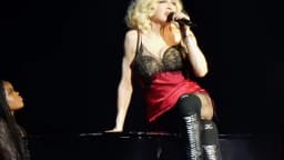 Madonna canta ao microfone durante apresentação da Celebration Tour, sentada sobre um piano no palco