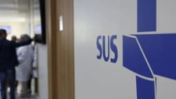 Corredor de unidade de saúde com o logo do SUS em destaque e profissionais de saúde ao fundo, foto ilustrativa