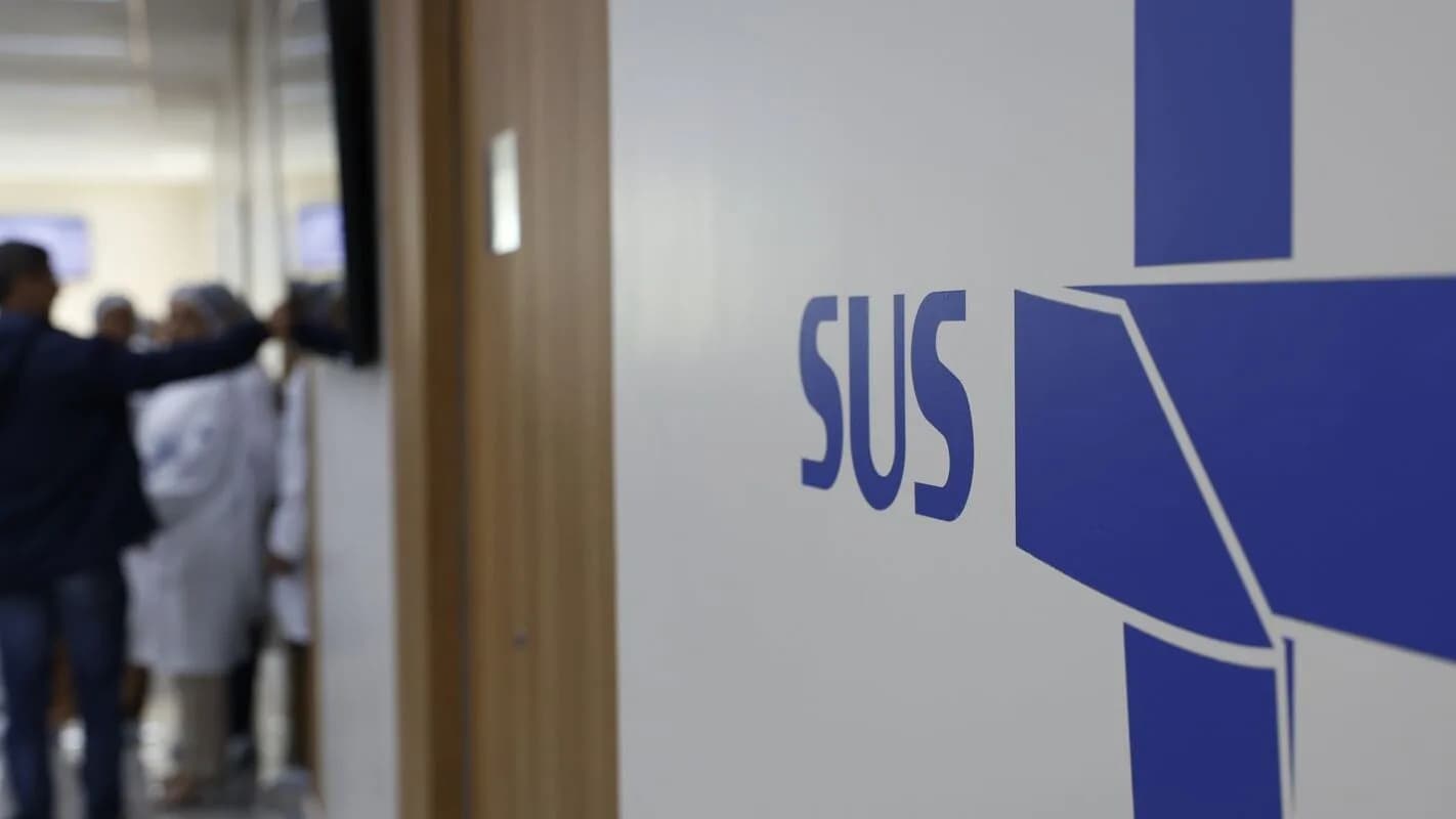 Corredor de unidade de saúde com o logo do SUS em destaque e profissionais de saúde ao fundo, foto ilustrativa