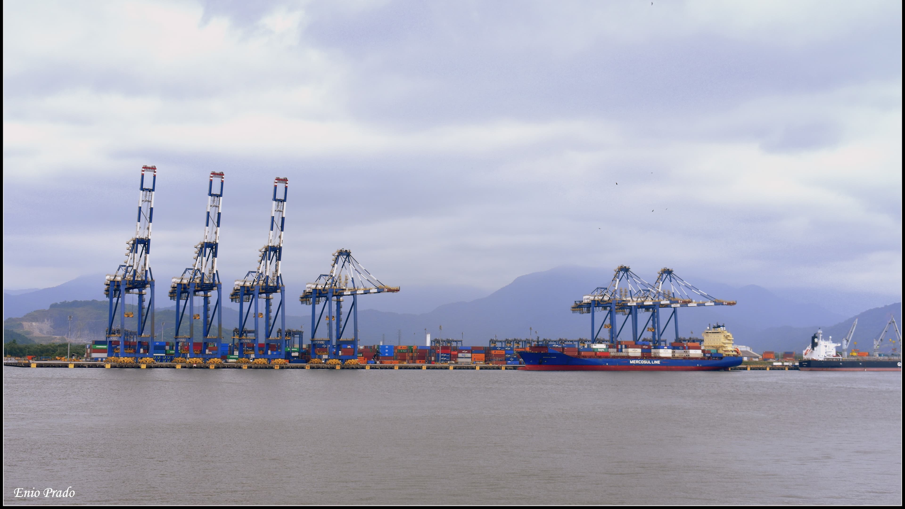 Guindastes portuários e navio cargueiro da Mercosul Line atracado no Porto de Santos, com contêineres empilhados no cais