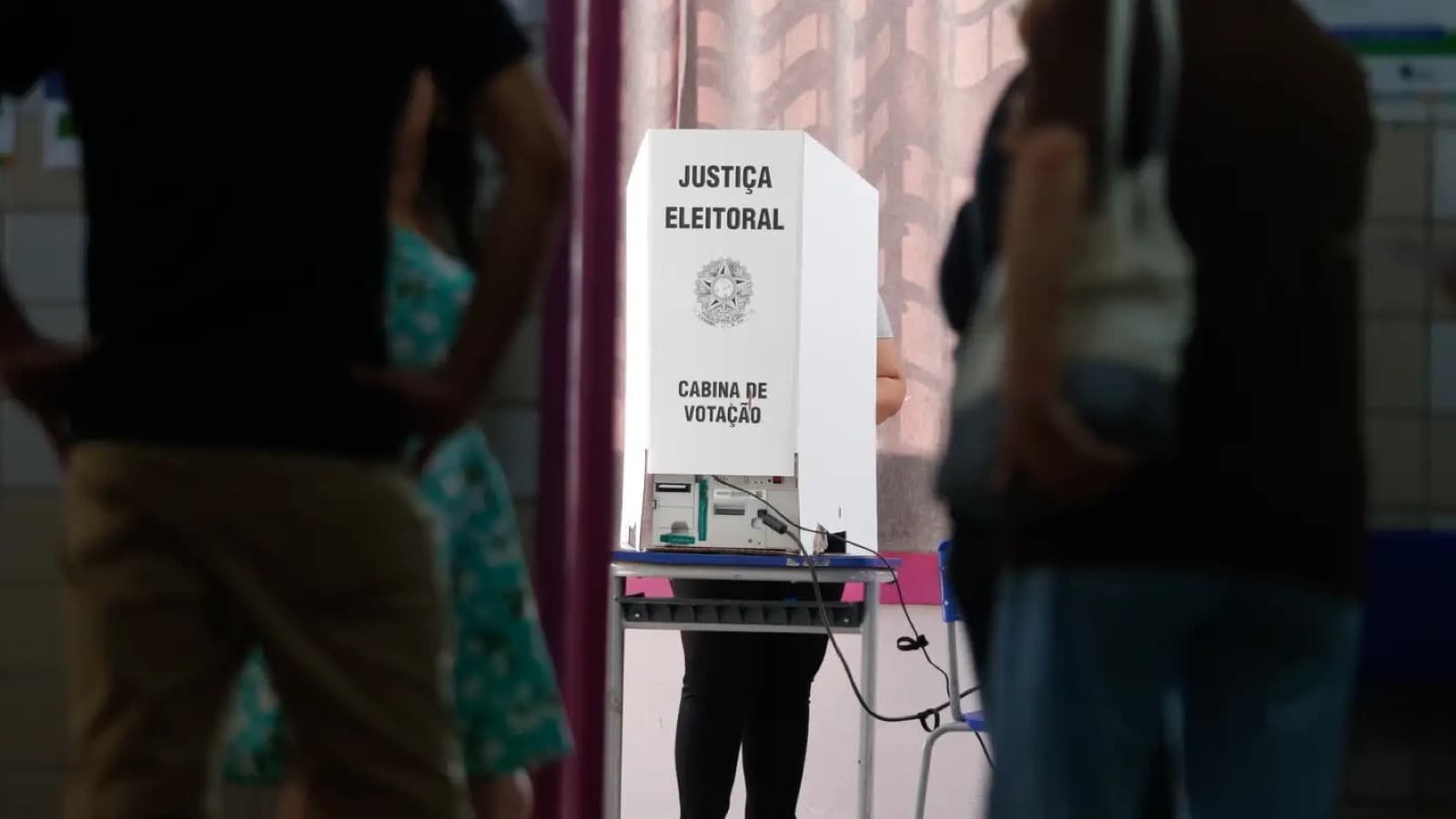 Eleitora vota em cabina da Justiça Eleitoral enquanto outras pessoas aguardam em pé na seção eleitoral; imagem de arquivo