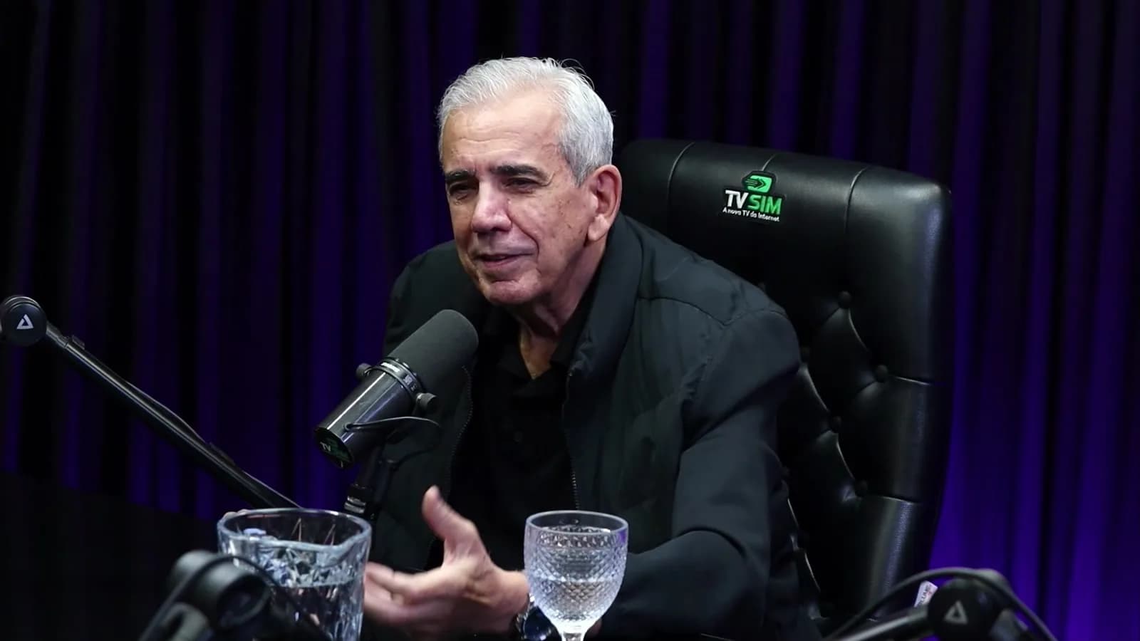 Ricardo Teagano fala gesticulando com a mão próxima ao microfone, durante entrevista em estúdio