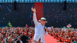 Lula de chapéu branco acena para multidão durante ato de campanha, com bandeira do Brasil ao fundo