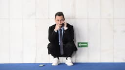 O senador Cleitinho aparece agachado, encostado em uma parede de mármore de um corredor do Senado, falando ao celular; um segundo aparelho está no chão, sobre o carpete azul. Imagem de arquivo.
