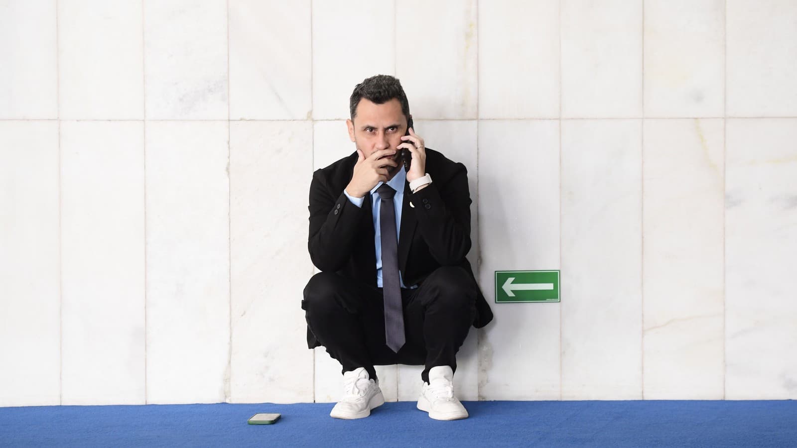 O senador Cleitinho aparece agachado, encostado em uma parede de mármore de um corredor do Senado, falando ao celular; um segundo aparelho está no chão, sobre o carpete azul. Imagem de arquivo.