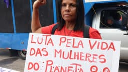 Manifestante de camiseta vermelha com o punho erguido segura cartaz pintado à mão com os dizeres Luta pela vida das mulheres do planeta durante ato de rua; imagem de arquivo