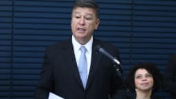 O senador Carlos Viana, de terno escuro, discursa ao microfone em evento de lançamento, com uma mulher sentada ao lado