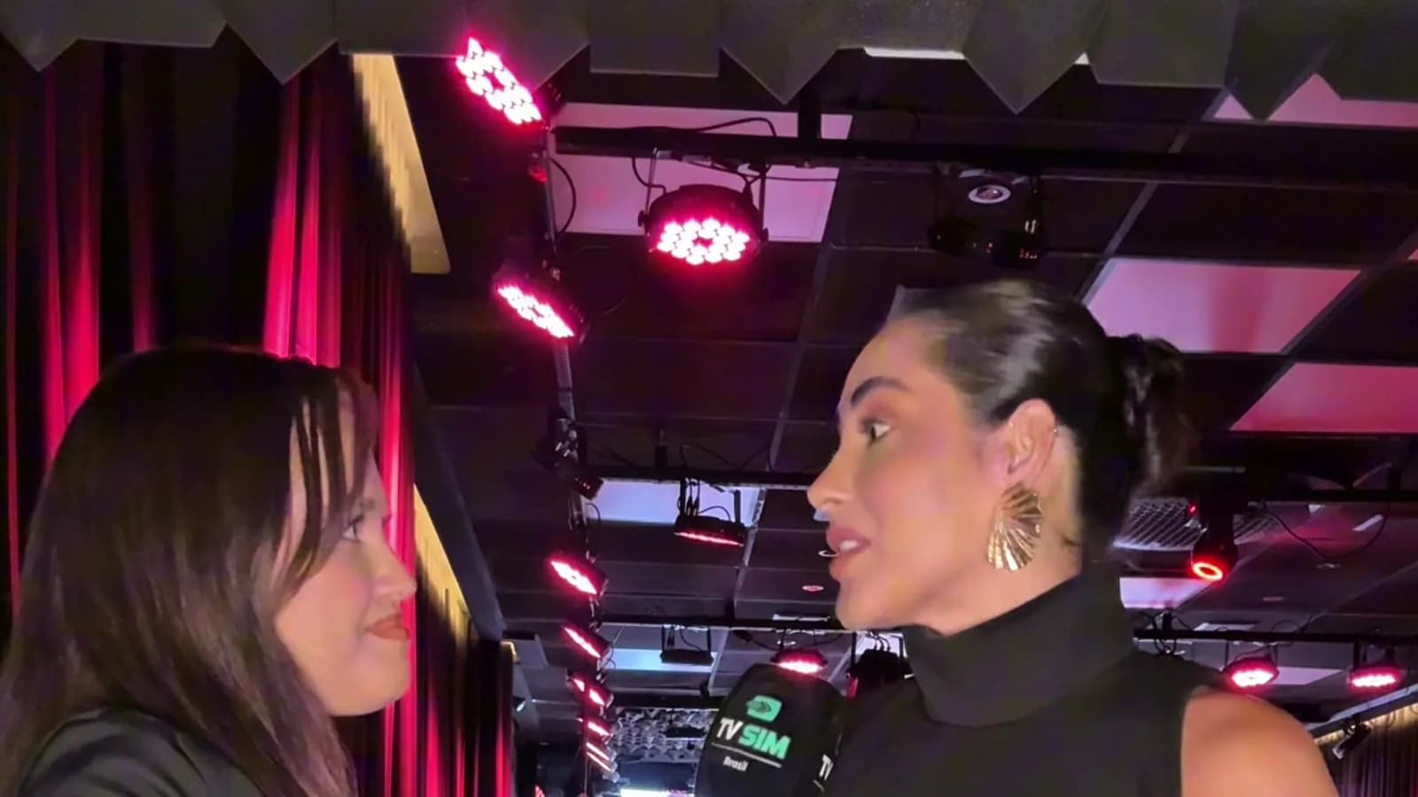 Repórter da TV Sim Brasil entrevista a influenciadora Anne, de macacão preto, durante evento com iluminação vermelha ao fundo