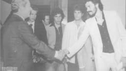 Fotografia em preto e branco de Sócrates, de bigode e barba, cumprimentando uma autoridade na década de 1980