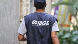 Agente do IBGE visto de costas, vestindo colete com a sigla do instituto, em atividade de campo